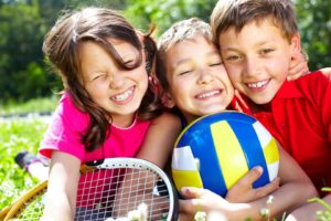 Three children with sports equipment embracing, looking at camera and smiling